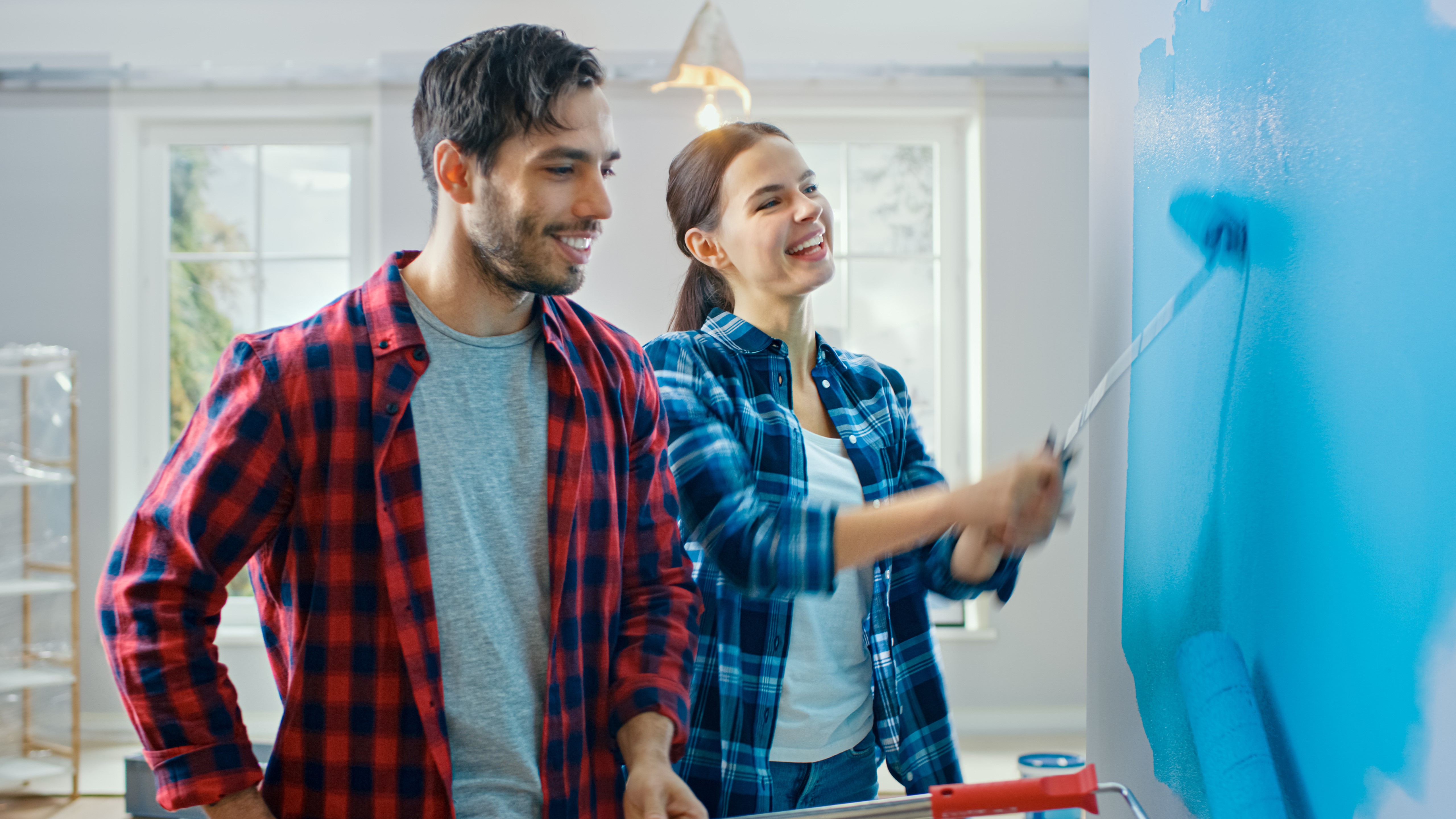 A person in a plaid shirt stands beside another person who is using a paint roller to apply bright blue paint to an interior wall in a well‑lit room with large windows and home furnishings in the background.